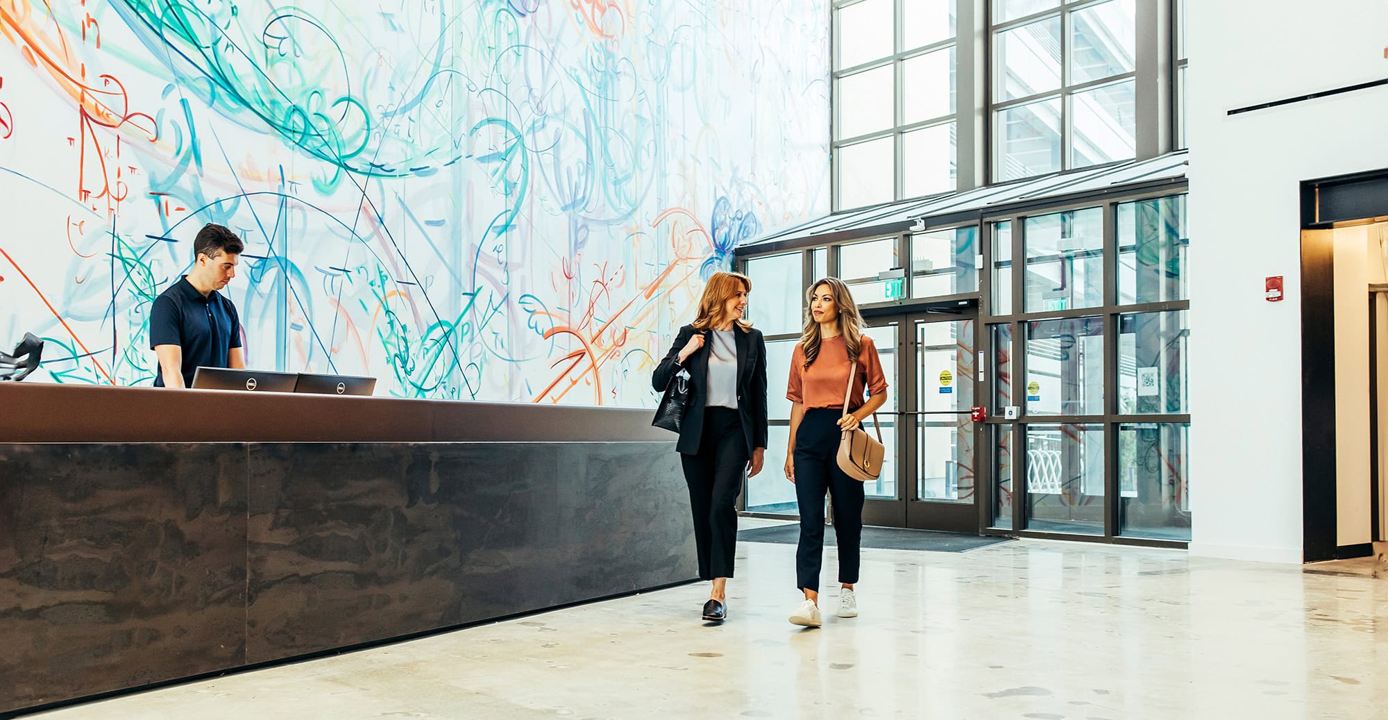 Two colleagues in a modern corporate lobby, chatting while walking past a mural.